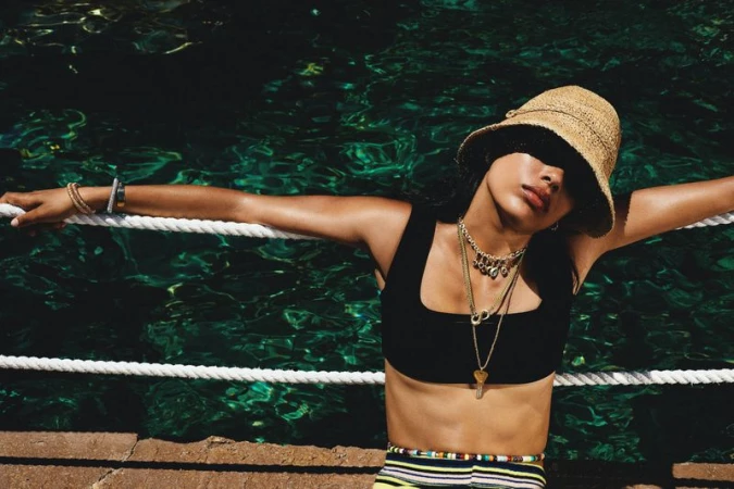 A woman in a straw hat poses near water, wearing a black top, striped skirt, and layered necklaces, leaning against a rope railing.