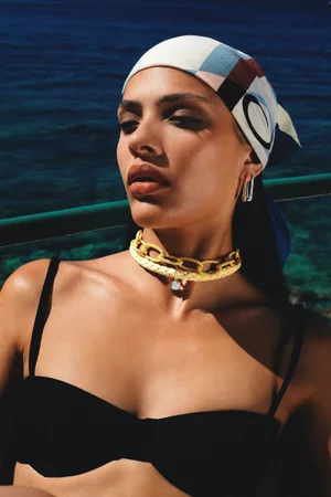Woman wearing a patterned headscarf, black bikini top, and yellow choker necklace against deep blue sea water.