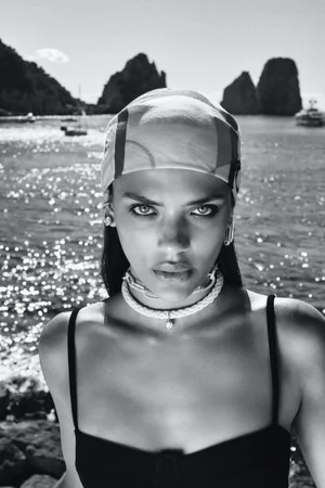 Black and white portrait of a woman wearing a headscarf, rope choker necklace, and black top against a sunlit coastal sea background.