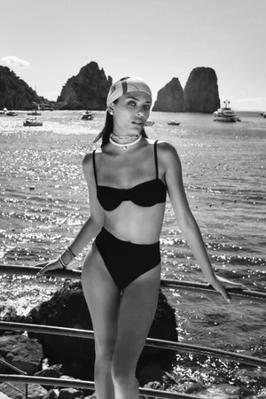 Monochrome fashion portrait of a woman in a black bikini and headscarf posing against coastal rock formations.