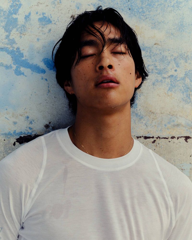 Portrait of a young adult male with dark hair and eyes closed, wearing a white long-sleeved shirt, against a mottled blue wall.