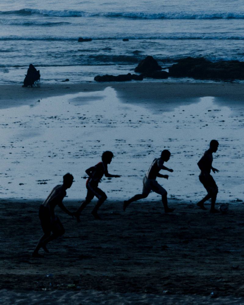 Silhouetted figures of boys playing soccer on a sandy beach with ocean waves in the background, captured with blue monochrome toning.