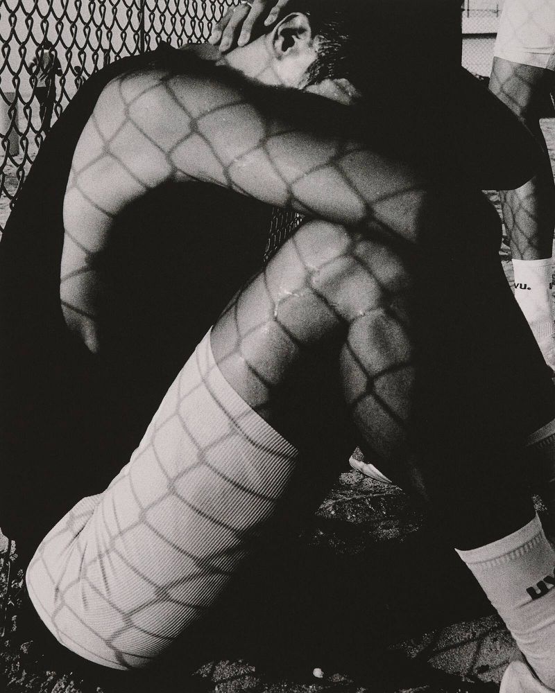Monochrome photo of a seated man with his head down, arms resting on his knees, and shadows from a chain-link fence on his body.
