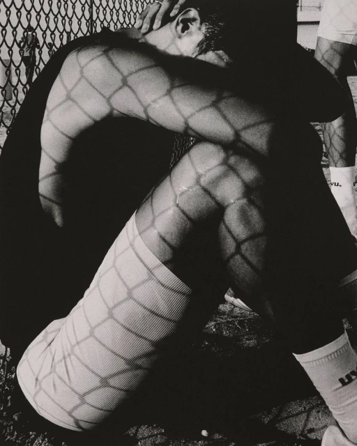 Monochrome photo of a seated man with his head down, arms resting on his knees, and shadows from a chain-link fence on his body.