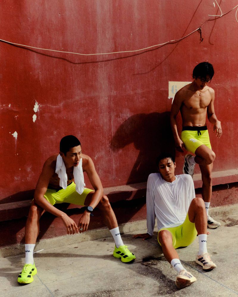 Three young adult men in sportswear posing outdoors against a red wall, showcasing fitness and athletic wear in bright sunlight.
