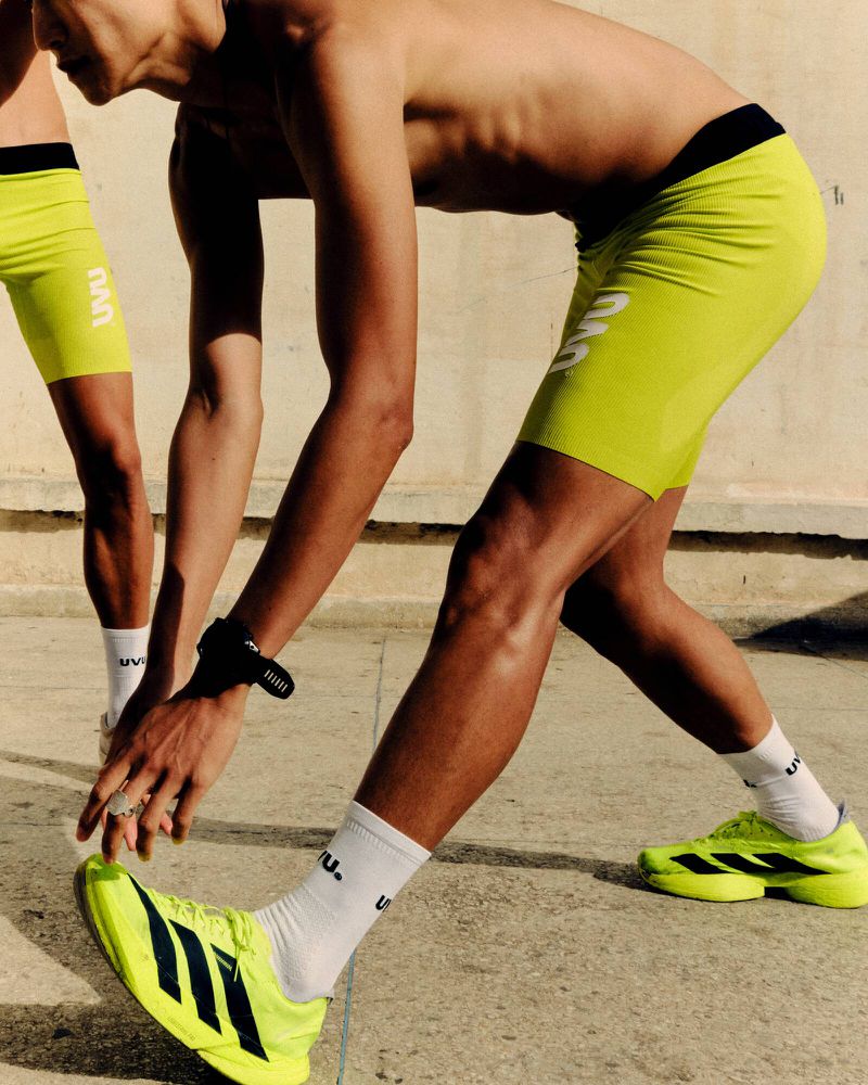 Two athletic men stretch outdoors wearing yellow shorts, socks, and running shoes, in a close-up shot emphasizing legs and feet.