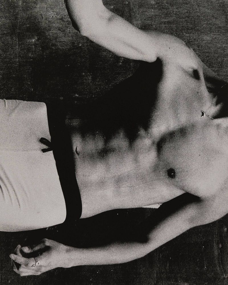 Monochrome photograph of a shirtless male torso and limbs against a dark background, showcasing light and shadow on skin and muscle.