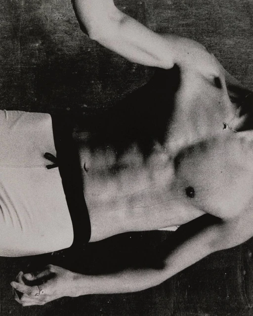 Monochrome photograph of a shirtless male torso and limbs against a dark background, showcasing light and shadow on skin and muscle.