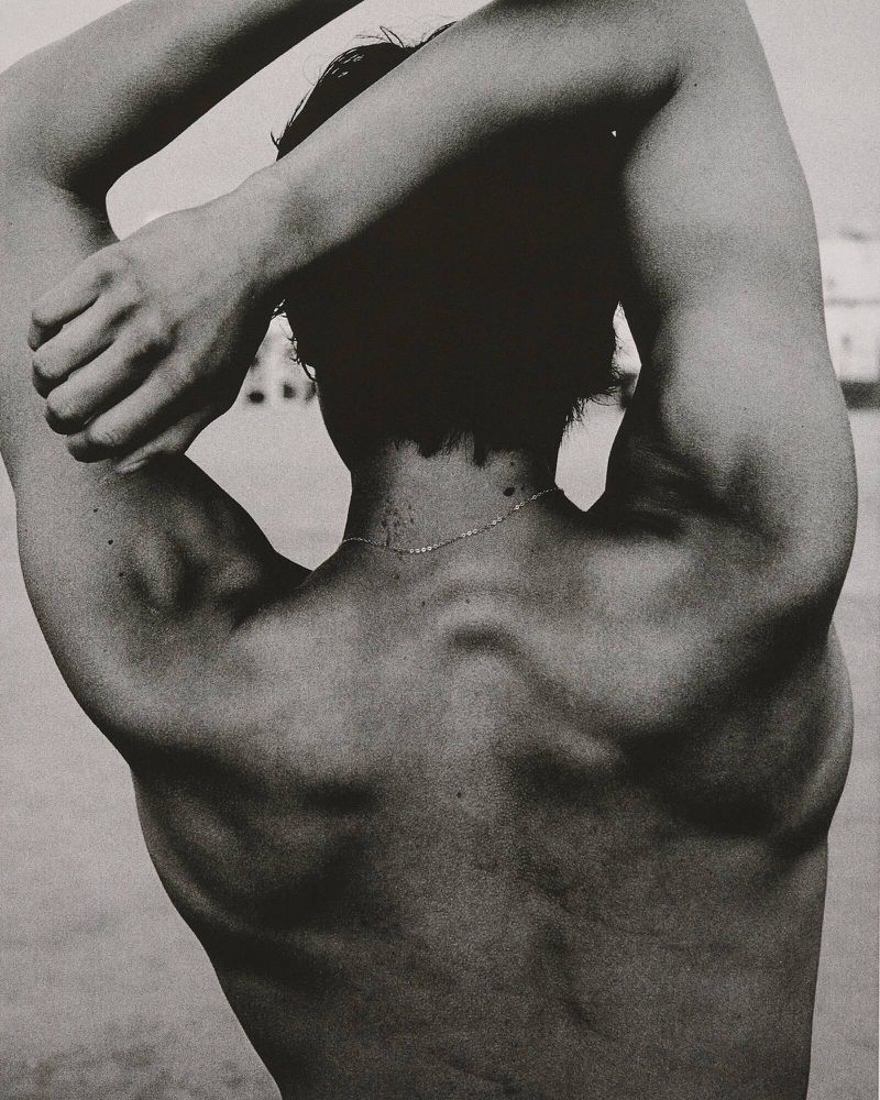 Black and white studio portrait of a shirtless adult male with arms raised behind his head, emphasizing musculature and physique.
