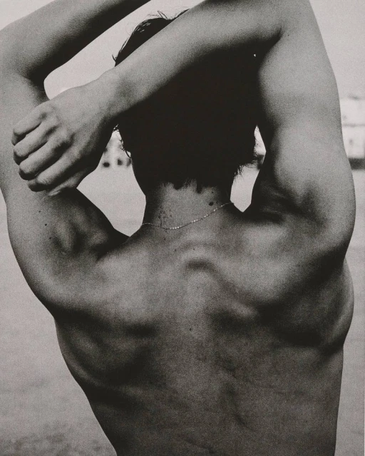 Black and white studio portrait of a shirtless adult male with arms raised behind his head, emphasizing musculature and physique.