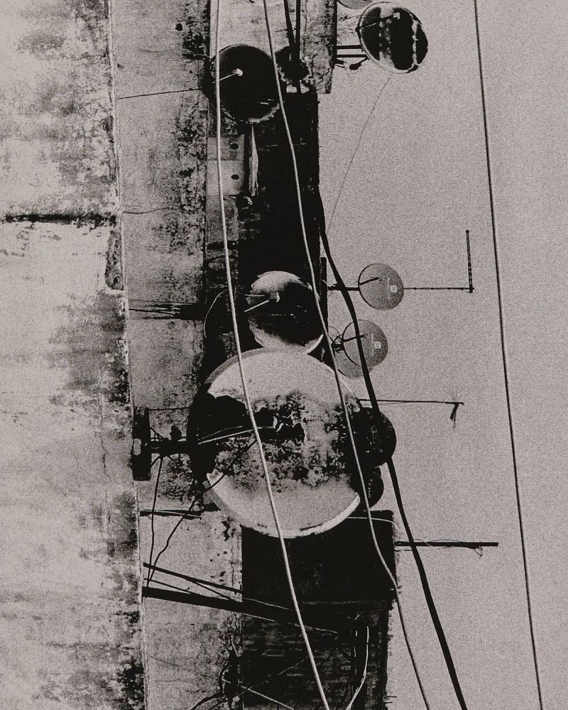 Abstract monochrome photograph of a weathered building with wires, poles, and round objects attached to a textured exterior surface.