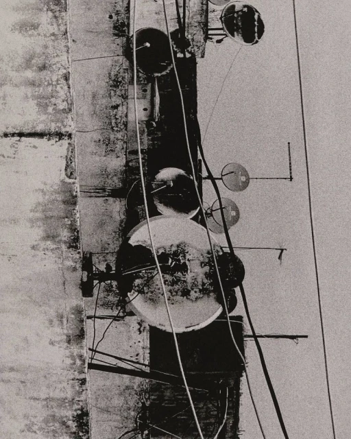 Abstract monochrome photograph of a weathered building with wires, poles, and round objects attached to a textured exterior surface.