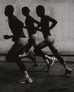 High-contrast monochrome shot of three male runners sprinting in athletic performance wear along a textured concrete wall.