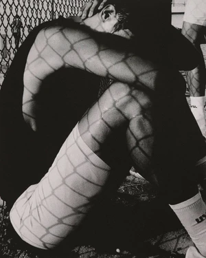 Black and white photo of a seated athlete with geometric chain link fence shadows across their arms, white athletic shorts, and socks.