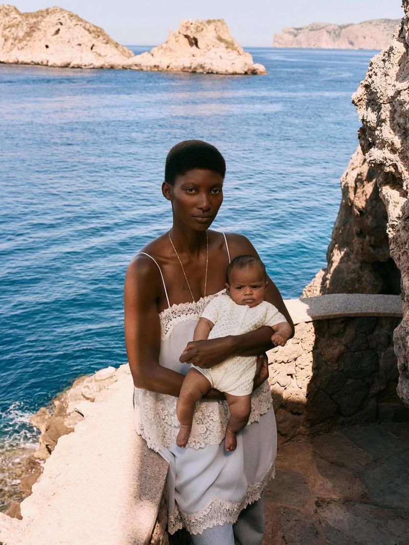 Model Mame Thiane Camara holding a baby in a knit romper on a stone balcony overlooking a rocky coastal sea.