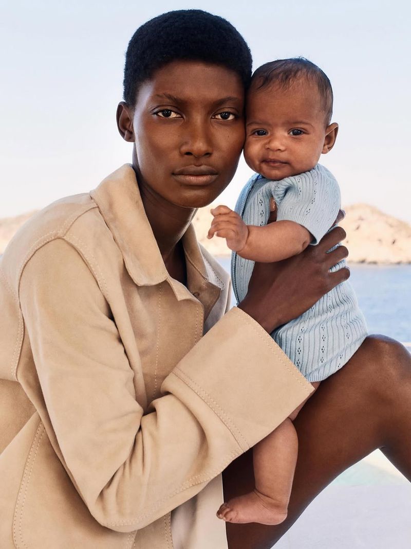 An elegant Black mother holds her mixed-race baby near a blurred ocean backdrop, both wearing light-colored clothing in a serene outdoor setting.