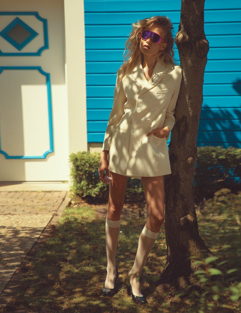 Stylish woman in a cream blazer and sunglasses leans against a tree in a sunny outdoor scene with blue door and lush greenery.