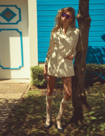 Stylish woman in a cream blazer and sunglasses leans against a tree in a sunny outdoor scene with blue door and lush greenery.