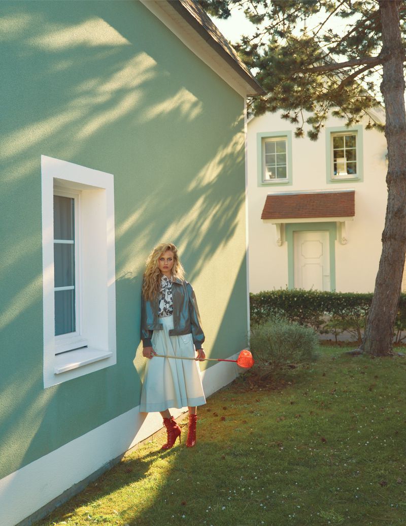 A fashionable adult female stands in a grassy yard wearing a jacket, blouse, skirt, and boots, holding a toy watering can. Buildings in background.