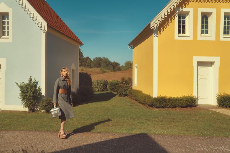 Fashionable woman in grey coat walking between bright yellow and blue buildings on a sunny day, holding metallic purse.