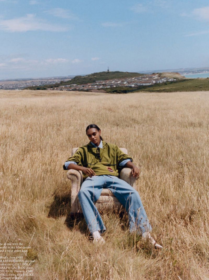 Man in blue jeans and a green jacket sits in an armchair in a golden field with a coastal cityscape in the background under a blue sky.