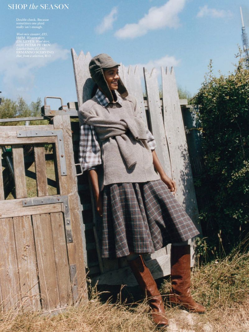 A smiling woman in a plaid skirt, vest, hat and boots stands near a weathered wooden gate against a blue sky in an outdoor setting.