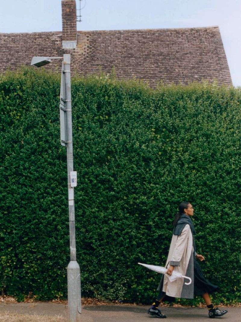 A woman in a trench coat walks past a green hedge, carrying a white umbrella. Streetlight and building in the background.