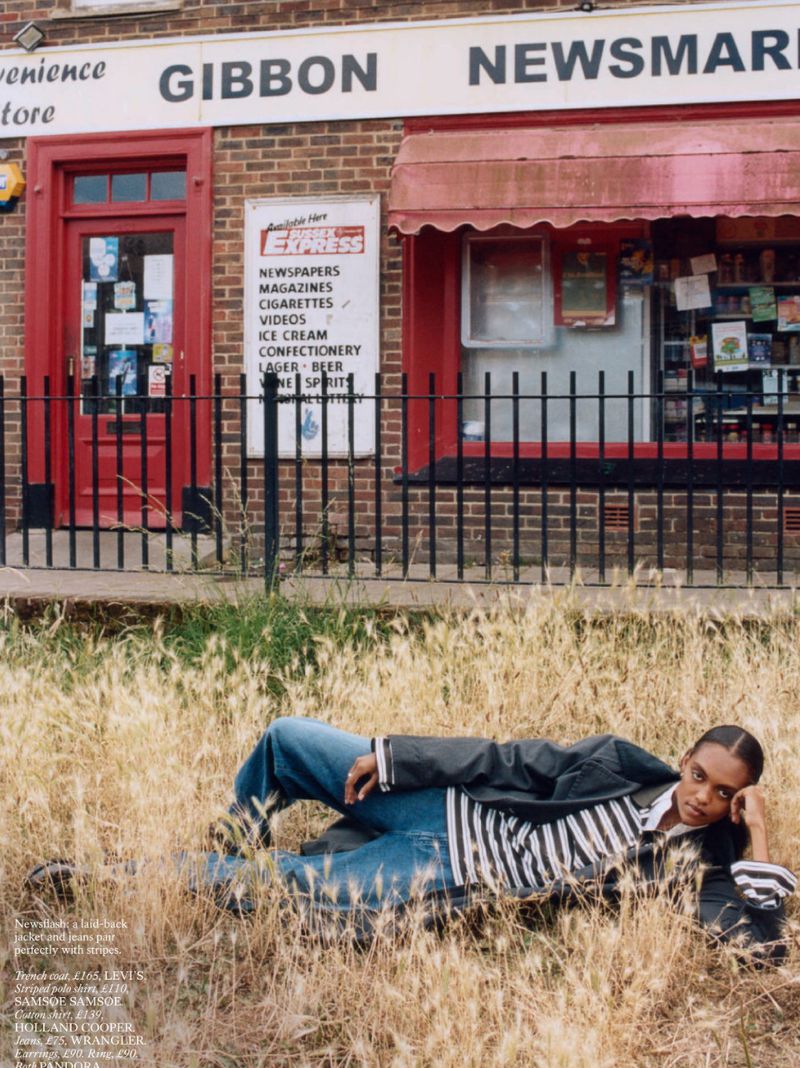 Adult woman lays in dry grass wearing denim pants, striped top, and black jacket in front of a brick convenience store. 