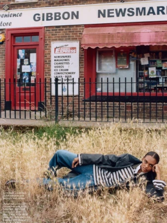 Adult woman lays in dry grass wearing denim pants, striped top, and black jacket in front of a brick convenience store. 