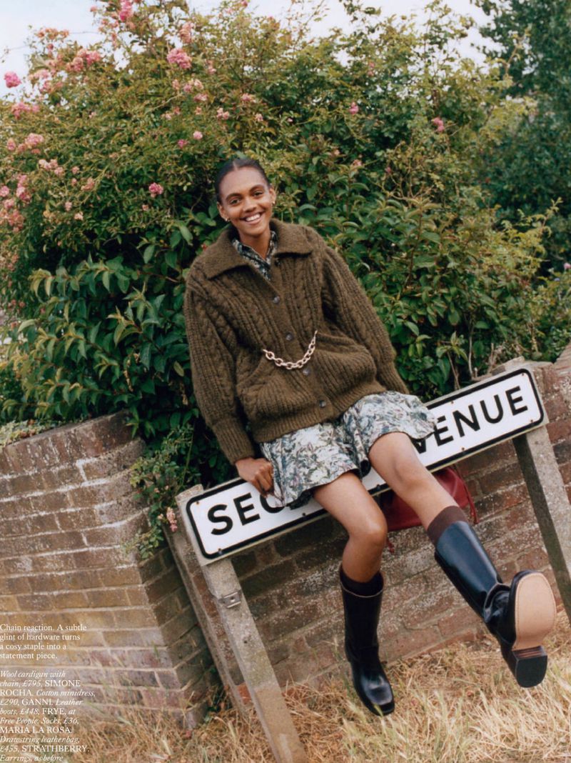 An adult female fashion model sits on a street sign in a rural setting, wearing a cardigan, dress, and boots, with a leafy background.