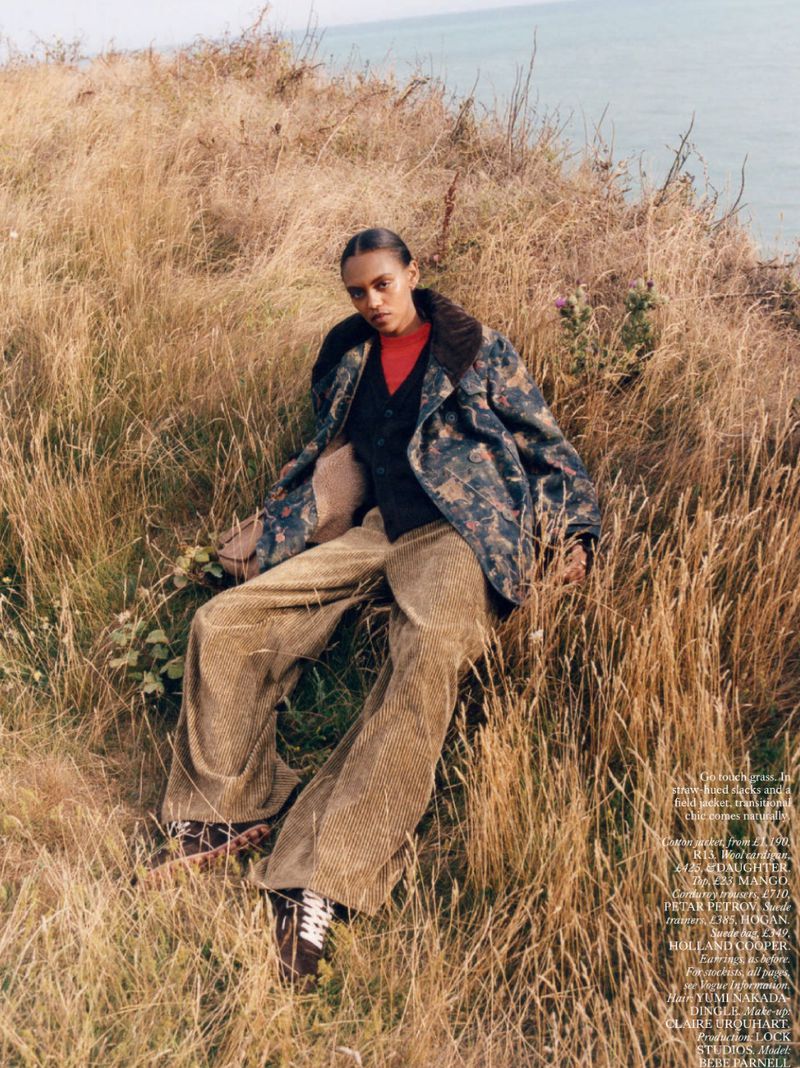 Woman in printed jacket and corduroy pants poses in a grassy field with ocean backdrop in outdoor fashion editorial shot.
