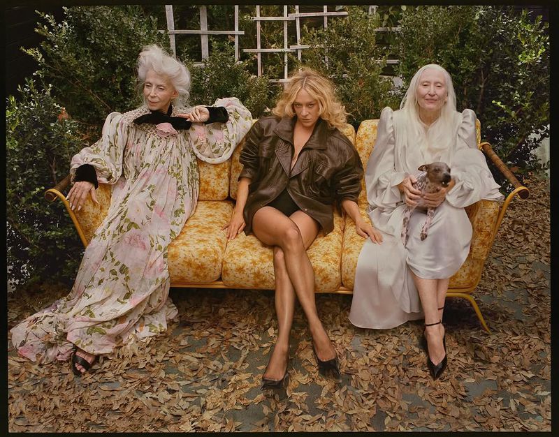 Three women pose on a yellow floral couch outdoors. Two elderly women sit on either side of a blonde woman wearing a jacket.