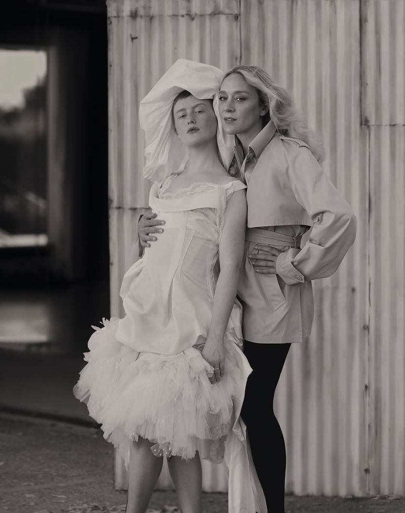 Two women pose together in a black and white fashion shot. One woman is wearing a tutu dress and the other a trench coat.