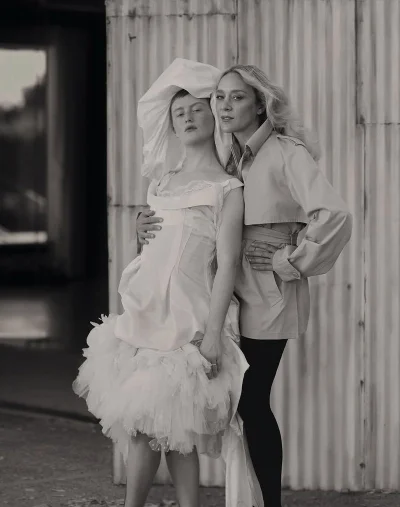 Chloe Sevigny in a belted trench coat standing beside a model in an avant-garde white tulle dress against a corrugated metal wall.