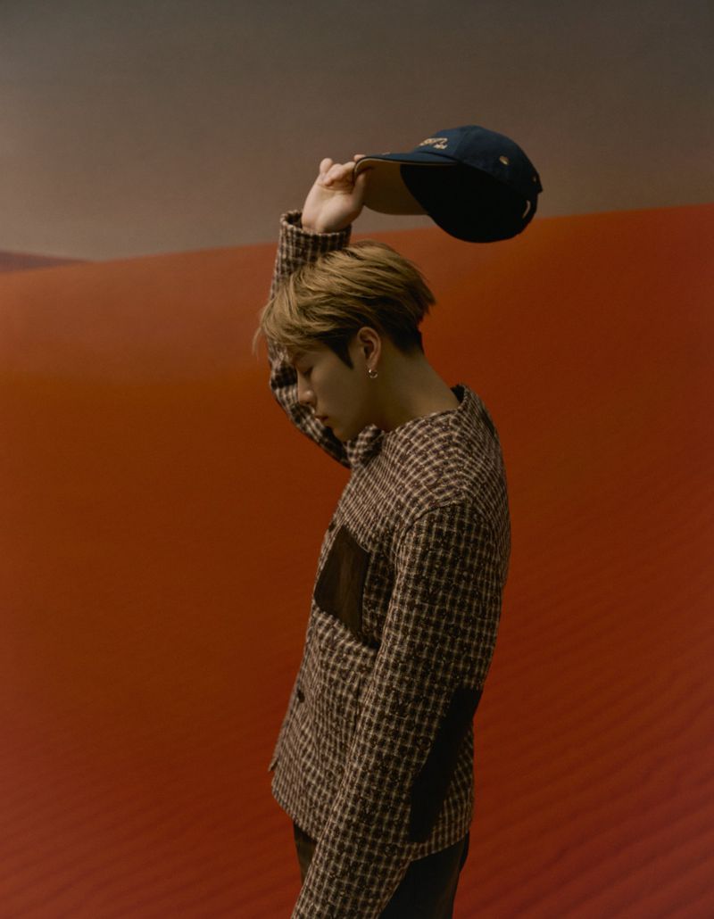 A person with blond hair in a patterned jacket stands indoors holding a baseball cap, with a red and grey blurred background.