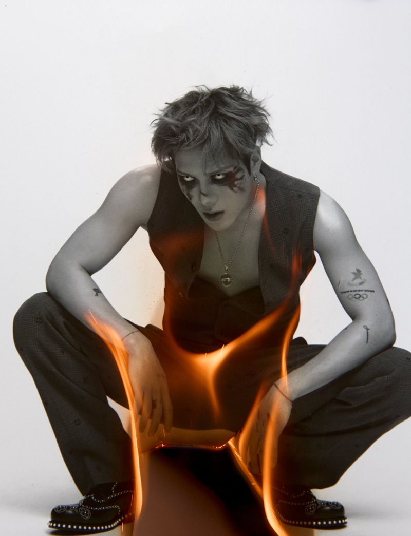 Young man with face makeup crouches in front of flames, wearing a vest and patterned pants in a white photo studio setting.