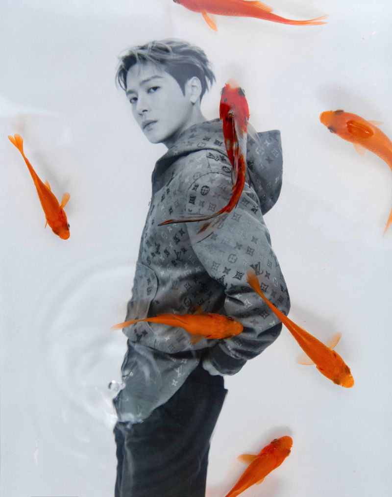 Monochrome portrait of a young adult male with short blond hair wearing a monogram hoodie surrounded by orange-colored fish.