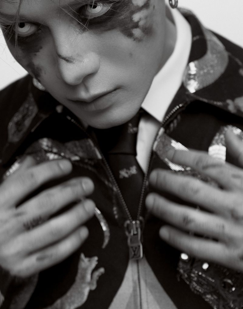 Monochromatic portrait of an adult with face paint and white contact lenses, wearing a patterned jacket and tie.
