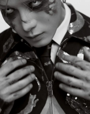 Monochromatic portrait of an adult with face paint and white contact lenses, wearing a patterned jacket and tie.