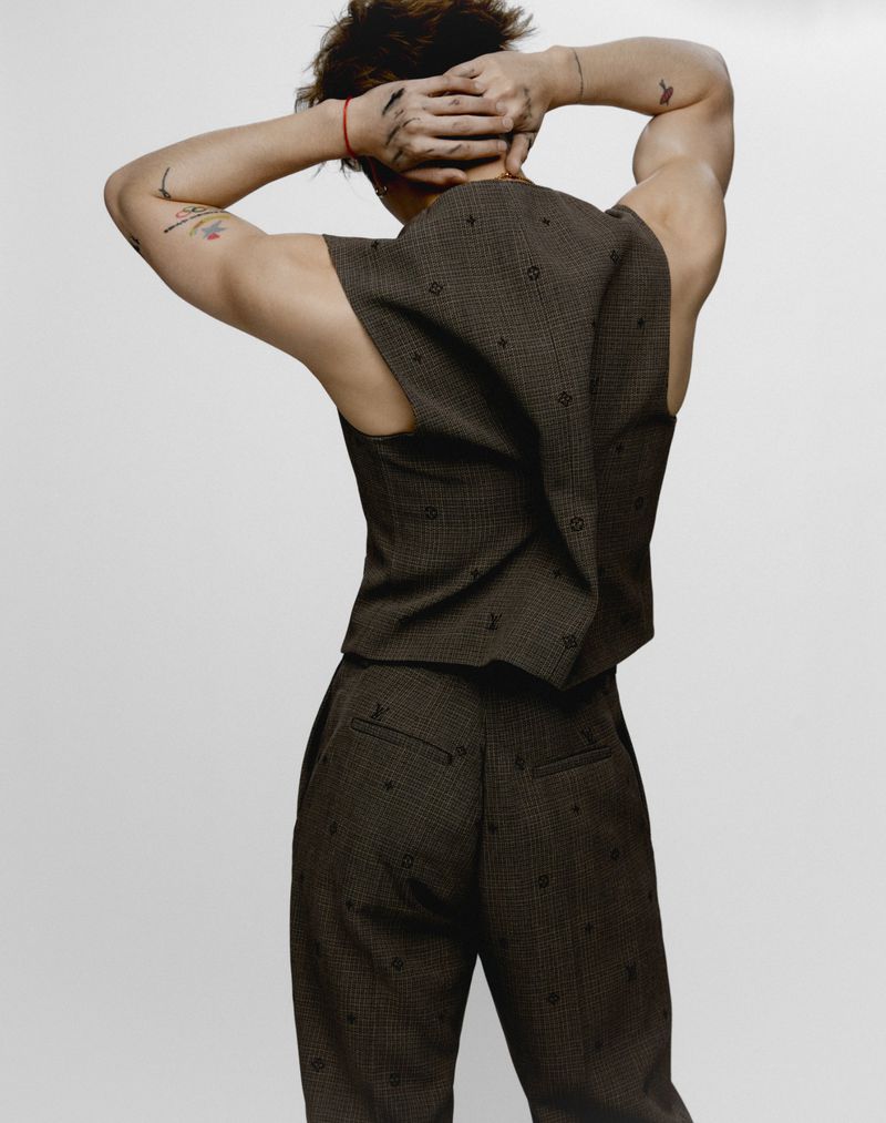 An adult male in a vest and trousers is seen from the rear. The man has his hands behind his head against a neutral background.