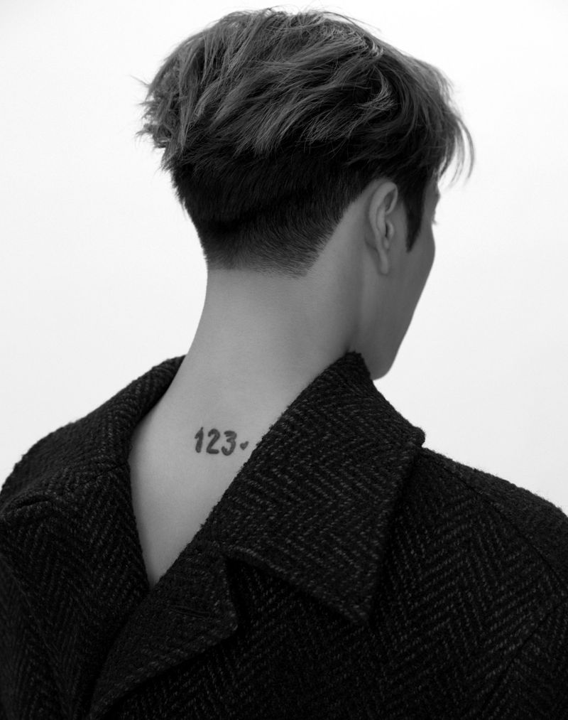 Monochromatic portrait of a person with dark hair and a herringbone jacket, revealing a tattoo on the back of their neck against a bright background.