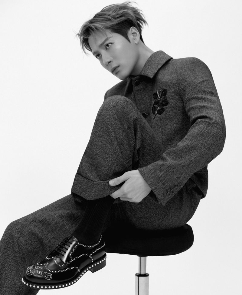 Monochrome studio portrait of a stylish man in a dark suit and embellished shoes, seated against a white background, captured in a fashion-forward pose.