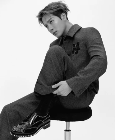 Monochrome studio portrait of a stylish man in a dark suit and embellished shoes, seated against a white background, captured in a fashion-forward pose.