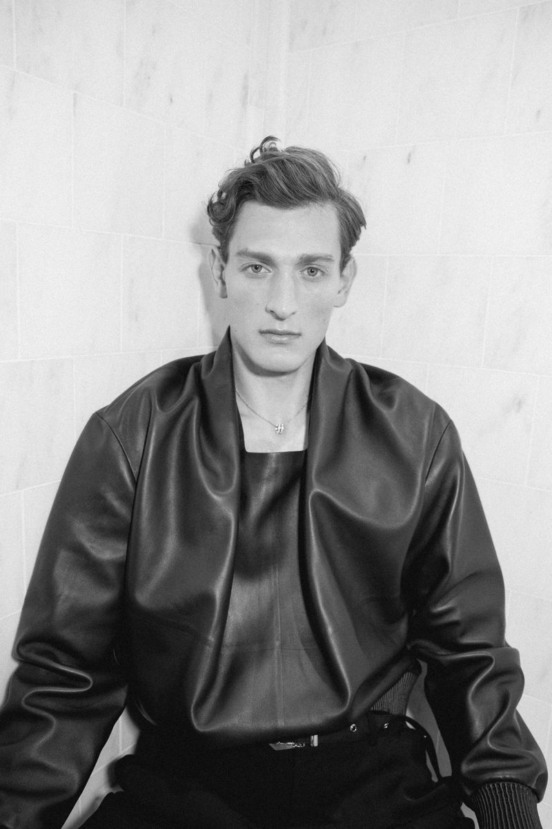 Monochrome portrait of a young adult male with dark hair wearing a black leather jacket against a neutral tile wall indoors.
