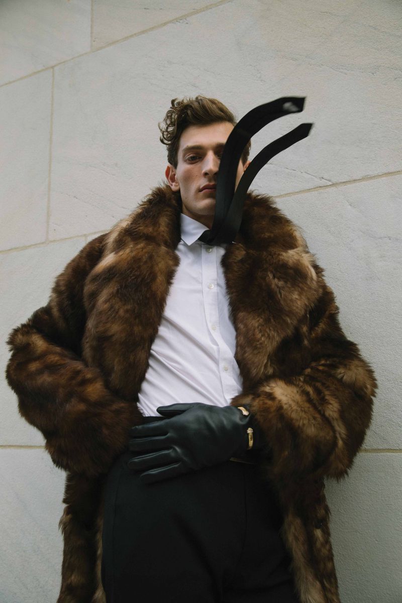 Fashionable man in fur coat poses against a wall with a necktie draped over his face, creating a striking editorial portrait.