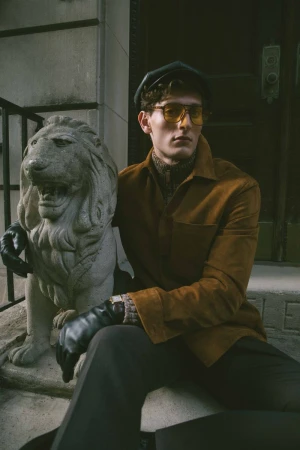 Stylish adult male model poses on stone steps next to lion statue wearing yellow sunglasses, suede jacket, and leather gloves outdoors.