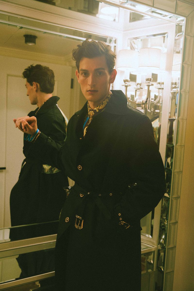 A fashionable man wearing a black trench coat poses in front of a mirror indoors, with a glimpse of a decorative lamp and wall behind him.