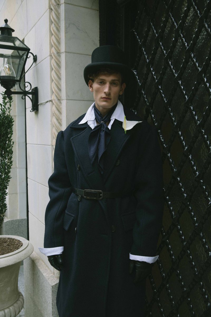 Man in black top hat and overcoat stands beside an ornate metal door. The image features a classic, somewhat formal fashion style.