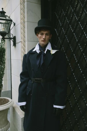 Man in black top hat and overcoat stands beside an ornate metal door. The image features a classic, somewhat formal fashion style.