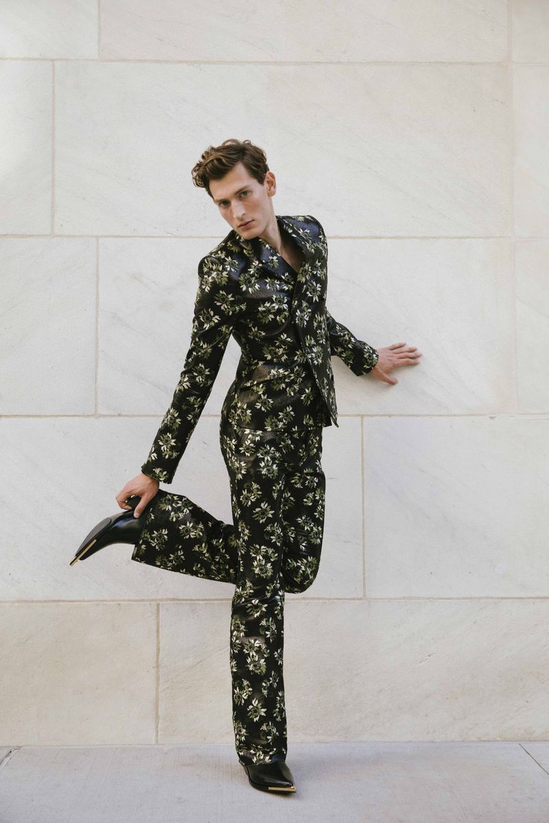 Male fashion model strikes a pose in a floral suit in front of a neutral wall, showcasing fashion style and visual appeal.